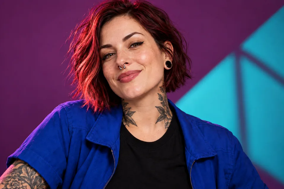 Model mit roten Haaren, Piercings und blauer Jacke vor violett-türkisem Hintergrund